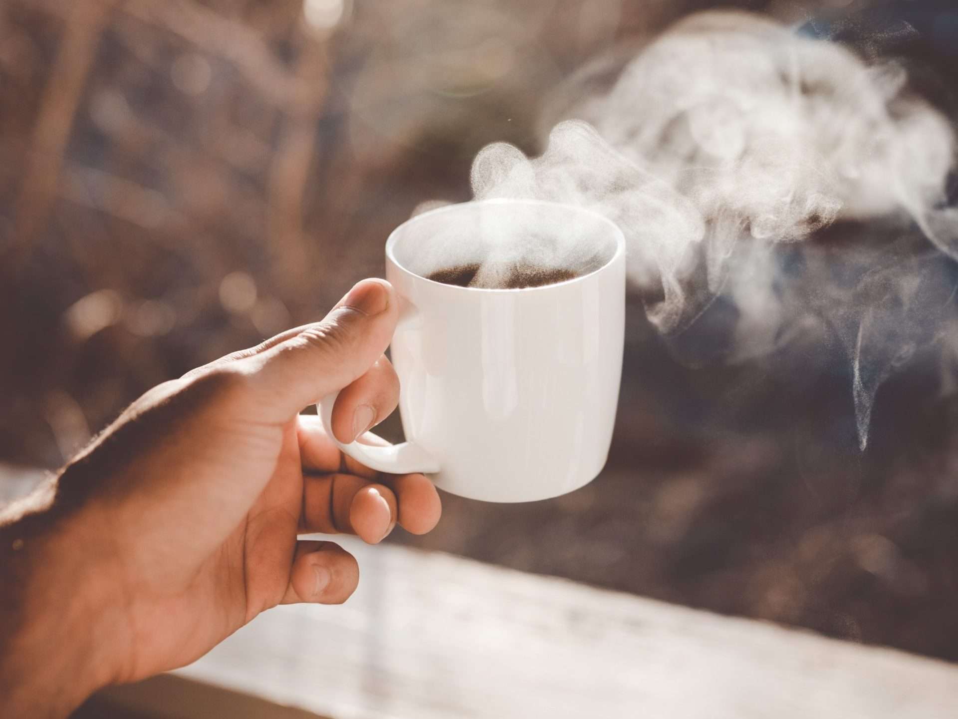 Hand holding a steaming cup of coffee in a sunlit environment.