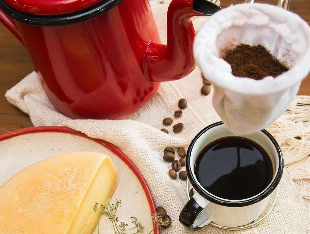 Cloth filter with coffee grounds hanging above a mug, with coffee beans and a red teapot in the background